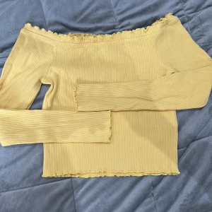 Gul ribbad offshoulder-top från Gina Tricot - Säljer en söt gul ribbad offshoulder-top från Gina Tricot. Toppen har långa ärmar och volangdetaljer vid halsringning och nederkant. Perfekt för dig som gillar en feminin och trendig stil!