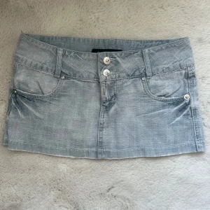 Ljusblå jeanskjol från Olala - Säljer en ljusblå jeanskjol från Olala med klassisk femficksdesign och snygga detaljer på bakfickorna. Kjolen har silverfärgade knappar framtill och är i en kort, lågmidjad modell som passar perfekt till sommaren.