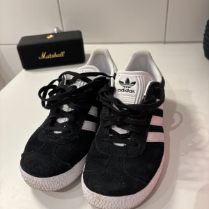 Svarta Adidas Gazelle sneakers - Säljer ett par klassiska svarta Adidas Gazelle sneakers med vita ränder och vit sula. Skorna har svart mocka, snörning och den ikoniska Gazelle-loggan i guld på sidan. Perfekta för dig som gillar stilrena och tidlösa sneakers. Dem är i storlek 36.💗💗💗