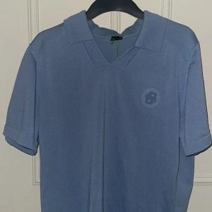 Ljusblå pikétröja från BOSS - Säljer en ljusblå pikétröja från BOSS med klassisk krage och korta ärmar. Tröjan har en diskret BOSS-logga och en rund badge med siffran 8 på bröstet. Perfekt för en stilren och sportig look.