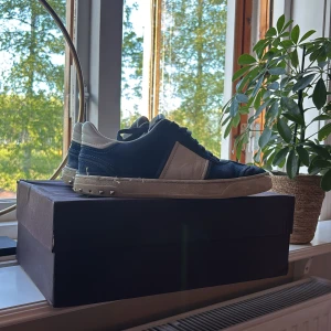 Blåa Valentino Garavani sneakers med vita detaljer - Säljer ett par blåa Valentino Garavani sneakers med vita detaljer och silverfärgade nitar vid hälen. Skorna har snörning och en platt sula. Kommer med originalkartong och dustbag.