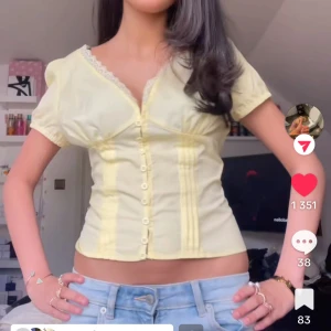 Gul blus med knappar och spetsdetalj - Söt ljusgul blus med korta puffärmar, knappar framtill och spetskant vid v-ringad hals. Figurnära passform och lätt croppad längd. Perfekt till jeans för en fräsch look.