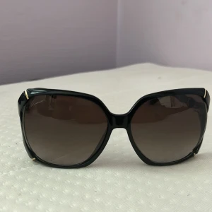 Svarta solglasögon från Gucci med gulddetaljer - Stilrena svarta solglasögon från Gucci med stora, fyrkantiga bågar och snygga gulddetaljer. Skalmarna har bambuinspirerad design och logotypen syns på sidorna. Mörka glas som ger en cool look.