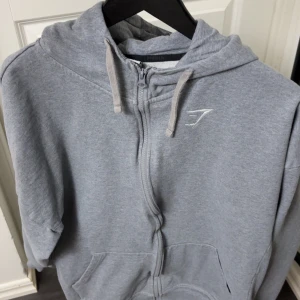Grå hoodie från Gymshark - Mjuk och bekväm grå hoodie från Gymshark med dragkedja framtill och huva med snören. Tröjan har fickor på magen och en diskret logga på bröstet. Perfekt för träning eller chill.