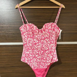 Rosa blommig baddräkt med smala axelband - En söt rosa baddräkt med vitt blommönster och smala axelband. Baddräkten har en hjärtformad urringning och är figurnära. Perfekt för sommardagar på stranden eller vid poolen.