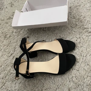 Svarta sandaler med klack och ankelrem - Säljer ett par stilrena svarta sandaler med blockklack och öppen tå. Skorna har en smal rem över tårna och en justerbar ankelrem med guldfärgat spänne. Perfekta för dig som vill ha en enkel men elegant look.