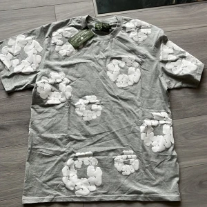 Grå t-shirt med vita blommor från Denim Tears - Säljer en grå t-shirt från Denim Tears med stora vita blommor tryckta över hela tröjan. T-shirten har rund hals och korta ärmar. Perfekt för dig som gillar streetwear och vill sticka ut med ett unikt mönster. Helt ny och är oanvänd. Jag säljer den för att jag inte gillade den