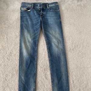 Blå jeans från Diesel - Säljer ett par klassiska blå jeans från Diesel med snygga slitningar och detaljerade sömmar. Jeansen har fem fickor och en liten Diesel-logga på myntfickan. Perfekta för dig som gillar en tidlös denimlook.