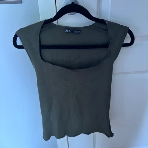 Olivgrön ärmlös topp från Zara - Säljer en olivgrön ärmlös topp från Zara med breda axelpartier och rundad halsringning. Toppen är croppad och har vågig nederkant, perfekt för en trendig look.