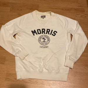 Vit sweatshirt från Morris - Vit sweatshirt från Morris med svart tryck framtill och rund hals. Klassisk modell med ribbade muddar vid ärmslut och nederkant. Perfekt för en avslappnad stil.