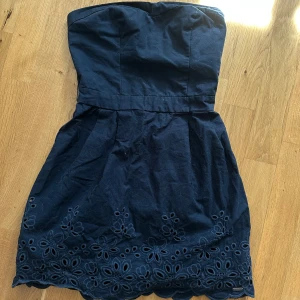 Sommarklänning från Hollister - Säljer en axelbandslös klänning från Hollister med broderade blommor och vågig nederkant. Klänningen har smock baktill för extra komfort och en feminin siluett. Perfekt för sommarens alla tillfällen! Svart/marinblå