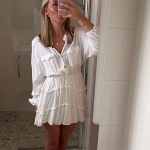 Vit volangklänning med knappar och knyt - Säljer en söt vit kortklänning med volanger och knyt i midjan. Klänningen har långa ärmar, bröstfickor och knappar framtill.