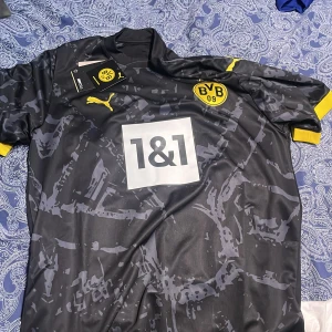 Borussia Dortmund fotbollströja - Säljer en svart Borussia Dortmund fotbollströja från Puma med gula detaljer och klubbmärke på bröstet. Tröjan har korta ärmar och ett mönster i grått samt sponsorlogga på framsidan. Helt ny köpte från Puma hemsidan!