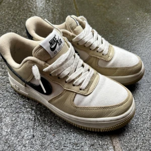 Nike Air Force 1 - Säljer ett par Nike Air Force 1 med beige och vita paneler samt svart swoosh. Skorna har klassisk snörning, rund tå och platt sula. Perfekta för dig som gillar stilrena sneakers med en twist. Originallåda finns.