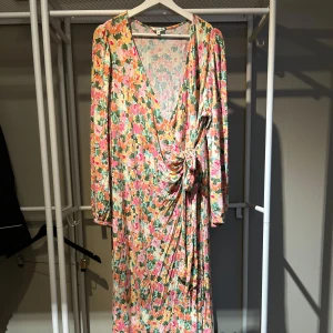Blommig omlottklänning med lång ärm - Säljer en långklänning med färgglatt blommönster i rosa, grön, gul och vit. Klänningen har omlottmodell med knyt i midjan, djup v-ringning och långa ärmar. Perfekt för dig som gillar färg och vill sticka ut. Det är fin ballongärm. Väldigt svalt skönt tyg men tungt fall. Är till golvet på mig som är 163! Passar xxl är från förra sommarens kollektion 