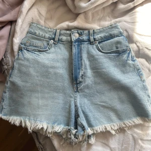 Ljusblå jeansshorts med fransar - Säljer ett par ljusblå jeansshorts med fransig kant nertill. Klassisk femficksmodell med knapp och dragkedja framtill. Perfekta för varma dagar och enkel att matcha med olika toppar. Oanvända💕