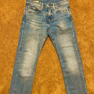 Levis 501 ljusblå jeans - Klassiska Levis 501 jeans i ljusblå tvätt med raka ben och femficksdesign. Byxorna har knappgylf och synliga slitningar för en vintagekänsla. Perfekta för dig som gillar tidlös denimstil.