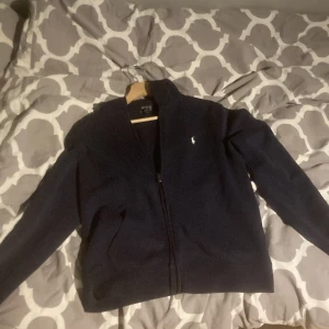 Mörkblå zip-hoodie från Polo Ralph Lauren - Mörkblå hoodie med dragkedja från Polo Ralph Lauren. Klassisk design med vit broderad logga på bröstet och två fickor framtill. Mjuk och bekväm, perfekt för vardagslooken. Tröjan är även i bra skick och perfekt för vardaglig användning.