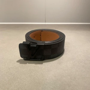 Svart LV bälte - Säljer ett svart bälte från Louis Vuitton med klassiskt rutigt mönster och stor LV-logga på spännet. Bältet har en stilren design med insida i brunt skinn och passar perfekt till jeans eller kostymbyxor.