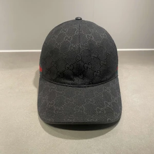 Svart gucci keps - Säljer en svart keps från Gucci med diskret GG-monogram över hela kepsen. Kepsen har en böjd skärm och klassisk passform samt grönt och rött band på sidan. Justerbar rem baktill för perfekt passform.
