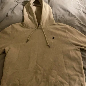 Beige hoodie med liten broderad logga - Säljer en beige hoodie med huva och dragsko. Tröjan har en liten broderad logga på bröstet och är tillverkad i mjukt material. Perfekt för en avslappnad stil.