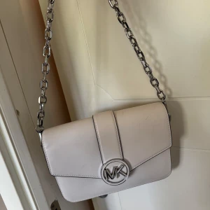 Vit axelväska från Michael Kors - Säljer nu min äkta Michael Kors väska, det är en stilren vit axelväska från med silverfärgad kedjerem och MK-logga framtill. Väskan har ett elegant och minimalistiskt utseende, perfekt för att lyfta din outfit. Inuti finns ett fack med dragkedja för småsaker. Kvitto finns kvar :) 