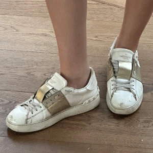 Valentino Garavani vita sneakers med gulddetaljer - Säljer ett par vita Valentino Garavani sneakers med  guldfärgad rem. Kommer med originalpåse och box. Storlek 38! Nypris ca 7200
