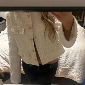 Elegant kofta  - Säljer en stilren vit tweed kavaj med fyra fickor framtill och dekorativa guldknappar. Jackan är croppad och har rund halsringning samt långa ärmar. Perfekt för att lyfta vilken outfit som helst.