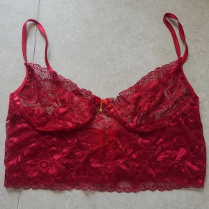 Röd spets bralette med rosett - Säljer en röd bhtopp i spets med tunna axelband och dekorativ rosett framtill. Toppen har blommigt spetsmönster och små gulddetaljer. Knäpps med hyskor i ryggen och har en croppad passform.