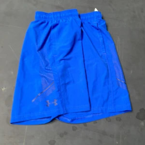 Blå träningsshorts från Under Armour - Snygga blå träningsshorts från Under Armour med elastisk midja och ventilerande meshpaneler på sidorna. Perfekta för gymmet eller löprundan. Klassisk logga på vänster ben och bekväm passform.