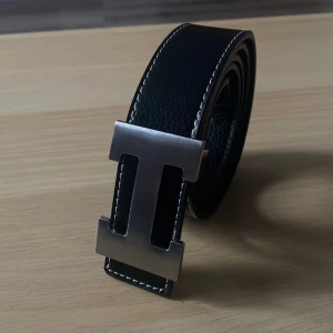 Svart skärp med H silverspänne - Stilrent svart bälte i skinn med ett stort, borstat silverspänne formad som ett H. Bältet har vita kontrastsömmar längs kanterna och passar perfekt till jeans eller kostymbyxor.