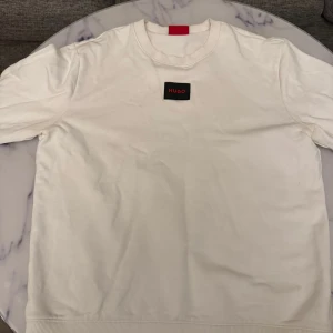 Vit sweatshirt från Hug0 - Säljer en stilren vit sweatshirt från Hug0 med diskret logga på bröstet. Tröjan har rund halsringning och långa ärmar med ribbade muddar. Perfekt för en clean och enkel look. Bra skick. Skickar alltid inom 24 timmar. 