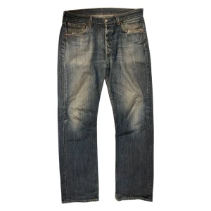 Levi's 501 blå jeans - Klassiska blå Levi's 501 jeans med rak passform och knappgylf. Jeansen har slitningar vid fickorna och en snygg tvättad look. Perfekta för en avslappnad stil och har den ikoniska röda Levi's-lappen bak.
