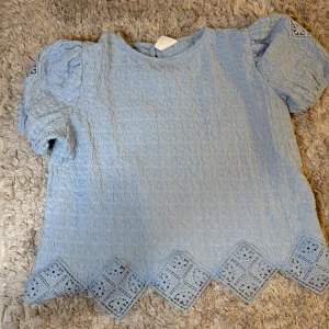 Ljusblå blus med spetsdetaljer - Supersöt ljusblå blus med puffärmar och dekorativa spetsdetaljer längs nederkanten. Blusen har rund halsringning och ett unikt mönster i tyget som ger en extra touch. Perfekt för dig som gillar romantisk stil.