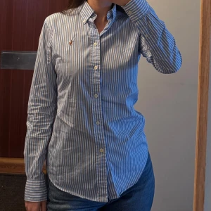 Blåvit randig skjorta från Ralph Lauren - Klassisk skjorta från Ralph Lauren i blått och vitt med smala ränder. Skjortan har lång ärm, knappar framtill och en liten broderad logga på bröstet. Perfekt till jeans eller chinos för en stilren look. Skjortan har ett hål vid armen (se bild) precis vid sömmen så går enkelt att sy. Jag kan inte sy därav säljer jag den. Storlek 4 står det på skjortan vilket motsvarar 36 eller small