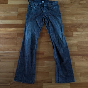 Mörkgråa bootcut jeans från Rock & Republic - Säljer ett par mörkgrå jeans från Rock & Republic med bootcut-passform. Jeansen har klassisk femficksdesign, snygga broderade detaljer på bakfickorna och stängs med dragkedja och knapp. Perfekta för dig som gillar en lite vidare stil nertill.