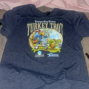Blå Turkey Trot 2019 t-shirt från PrintLase - Grå t-shirt  med stort tryck fram och bak från Turkey Trot 2019. Motivet fram visar en tecknad kalkon och sköldpadda som springer. T-shirten har rund hals och är tillverkad i mjuk polyesterblandning.