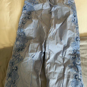 Blåvitrandiga broderade byxor med dragsko - Säljer ett par blåvitrandiga byxor med bred passform och fina broderade detaljer längs benen. Byxorna har dragsko i midjan och är perfekta för en avslappnad men ändå stilren look. Aldrig använda, från chloe schutermanns kollektion