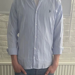  Polo Ralph Lauren skjorta L - Säljer en ljusblå skjorta från Polo Ralph Lauren i slim fit-modell. Skjortan har långa ärmar, klassisk krage och knäppning framtill. Broderad logga på bröstet. Perfekt för en stilren look! Storlek L