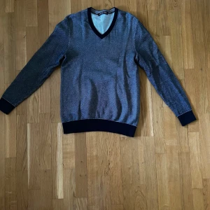 Grå v-ringad tröja från Michael Kors - Säljer en stilren grå v-ringad tröja från Michael Kors. Tröjan har mörkblåa ribbade muddar vid ärmslut, nederkant och runt halsen. Perfekt för dig som gillar klassisk och enkel stil.