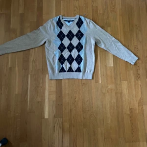 Beige argyletröja från Tommy Hilfiger - Snygg beige tröja från Tommy Hilfiger med klassiskt argylemönster i mörkblått och grått framtill. V-ringad modell med långa ärmar, perfekt för lager-på-lager. Liten broderad logga på bröstet.