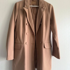 Beige kavaj  - Stilren beige kappa från Ambiu med dubbelknäppning och klassisk krage. Jackan har långa ärmar och två fickor framtill. Perfekt för dig som vill ha en tidlös och elegant look.