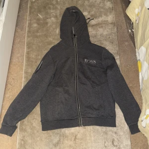 Mörkgrå huvtröja från Boss - Säljer en mörkgrå huvtröja från Boss med dragkedja framtill. Tröjan har en justerbar huva och ribbade muddar vid ärmslut och nederkant. Perfekt för en avslappnad stil. Storlek är M men en person som har storlek S kan också ta på sig den.