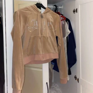 Beige hoodie från Juicy Couture - Säljer en snygg beige hoodie från Juicy Couture med glittrande detaljer på bröstet. Den har en dragkedja framtill och en bekväm huva. Perfekt för en avslappnad stil.