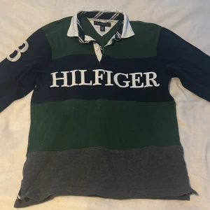 Randig pikétröja från Tommy Hilfiger - Säljer en stilren randig pikétröja från Tommy Hilfiger i grönt, blått och grått. Tröjan har långa ärmar och en klassisk krage med knappar. Perfekt för en avslappnad look.