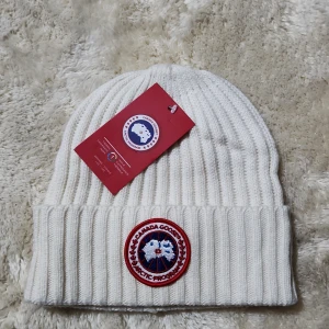 Vit ribbad mössa med märke - Snygg vit ribbad mössa med ett broderat märke framtill. Tillverkad av 64% bomull och 36% polyester, perfekt för kyliga dagar. Mössan har en klassisk design med uppvikt kant.