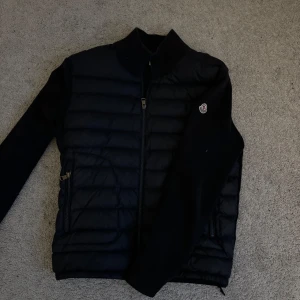 Svart pufferjacka från Moncler - Snygg svart pufferjacka från Moncler med dragkedja och broderad logga på bröstet. Jackan har en stilren design med långa ärmar och ribbad krage. Perfekt för kyligare dagar.