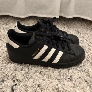 Svarta Adidas Superstar sneakers - Säljer ett par klassiska svarta Adidas Superstar sneakers med vita ränder och snörning. Skorna har den ikoniska tåhättan och Adidas-loggan på hälen. Perfekta för en stilren look.