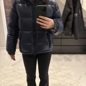 Mörkblå pufferjacka från Lexington - Säljer en snygg mörkblå pufferjacka från Lexington. Jackan har en dragkedja framtill och en mysig huva. Perfekt för kyliga dagar med sin vadderade design. Den är både stilren och funktionell. Helt nya använd fåtal gånger. Finns liten skada på jackan vid knapparna  nere som spänner till jackan. Bild kan skickas via dm om man vill se. Sitter som en s i storlek passar både folk i xs och s.