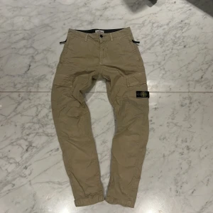 Beige cargobyxor från Stone Island - Snygga beige cargobyxor från Stone Island med klassisk logga på benet. Byxorna har flera praktiska fickor och en bekväm passform. Perfekta för en avslappnad stil. Pris kan lätt diskuteras vid snabb affär. 